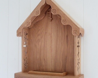 May include: A light brown wooden wall shelf featuring a gabled roof design with scalloped edges and decorative perforations. The side panels display intricate laser-cut cross and leaf motifs, while a small rectangular tray sits on the base.