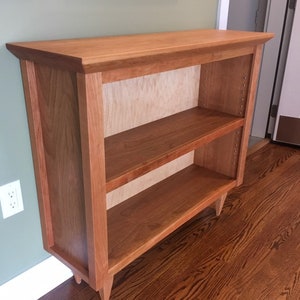 May include: A wooden bookcase with two shelves and a light wood finish. The bookcase has a simple design with a slightly angled base.