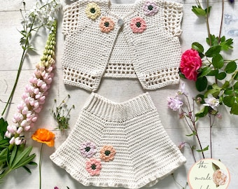 May include: A white crocheted baby outfit with a cardigan and skirt. The cardigan has a button closure and both pieces are decorated with colorful crocheted flowers.