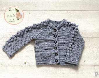May include: A gray crocheted cardigan with a button closure and pom-poms on the sleeves. The cardigan is laid flat on a light wood surface. The text "The moule hole" is visible in the upper left corner of the image.