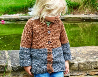 May include: A color-blocked crochet cardigan featuring a brown upper section and a dark gray lower section with a speckled texture. The garment has long sleeves, a ribbed hem, and a vertical front placket with five dark buttons. Blue denim jeans.