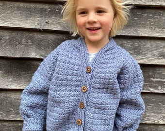 May include: A child wearing a blue crocheted cardigan with wooden buttons, a brown beanie, and blue jeans. The cardigan has a textured stitch pattern. The child is standing in front of a wooden wall with a circular sticker that says "The moule hole".