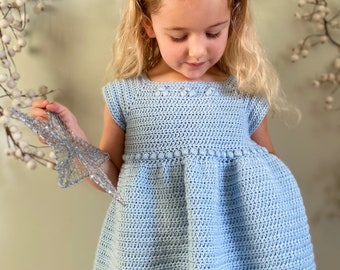 May include: A little girl wearing a light blue crochet dress with a star headband. The dress has a scalloped hem and a crocheted flower detail on the bodice. The girl is holding a silver star.