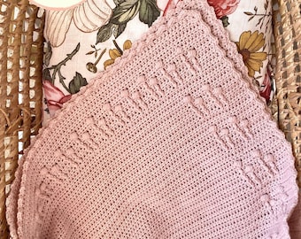 May include: A pink crocheted baby blanket with a scalloped edge and a textured pattern. The blanket is resting on a floral patterned pillow inside a wicker basket.