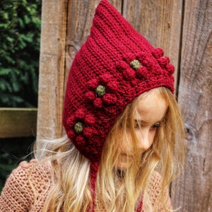 Può includere: Un cappello rosso all'uncinetto con decorazioni di fiori verdi. Il cappello ha una corona a punta e una lunga cravatta.