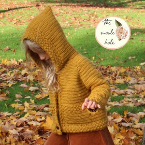 Op de afbeelding: Een jong persoon draagt een mosterdgele gebreide hoodie met een bruine corduroy rok en zwarte laarzen. De persoon staat in een stapel herfstbladeren. De afbeelding bevat de tekst "The moule hole".