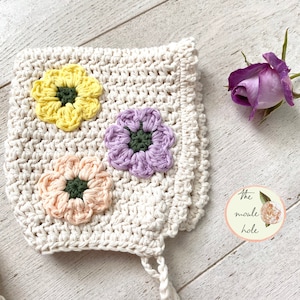 Könnte beinhalten: Eine gehäkelte Babyhaube mit weißem Hintergrund und drei gehäkelten Blumen in Gelb, Lila und Pfirsich. Die Blumen haben grüne Zentren. Die Haube hat eine Bindekordel. Der Text "The noodle hole" befindet sich auf einem gelben Kreis mit einer rosa Blume.