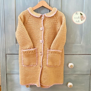 Könnte beinhalten: Ein langer, gehäkelter Cardigan in Senfgelb mit rosa Besatz. Der Cardigan hat einen Kragen, einen Knopfverschluss und zwei Taschen.