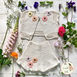 May include: A white crocheted baby outfit with a cardigan and skirt. The cardigan has a button closure and both pieces are decorated with colorful crocheted flowers.