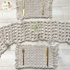 May include: Three crocheted placemats with fringe edges. The placemats are made of natural beige cotton rope and have a square pattern. The placemats are arranged on a light wood surface with gold silverware and a glass.