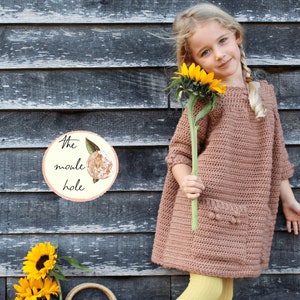 May include: A young girl wearing a brown crocheted dress, yellow tights, and brown boots holds a sunflower. She is standing in front of a wooden wall with a sign that reads "The moule hole".