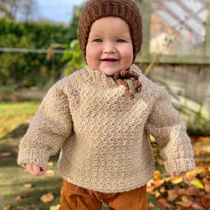 May include: A toddler wearing a brown crocheted sweater and matching hat, brown corduroy trousers, and white socks. The toddler is standing on a wooden surface with fallen leaves. The text "The moule hole" is visible in the lower right corner of the image.