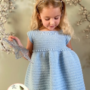 Può includere: Una bambina che indossa un abito di uncinetto azzurro chiaro con una fascia per capelli a forma di stella. L'abito ha un orlo a balze e un dettaglio floreale lavorato a uncinetto sul corpetto. La bambina tiene una stella d'argento.