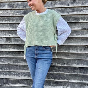 May include: A light green crochet vest with side ties, worn over a white button-down shirt and paired with blue jeans. The vest has a textured, open weave design. The jeans are a classic blue wash.