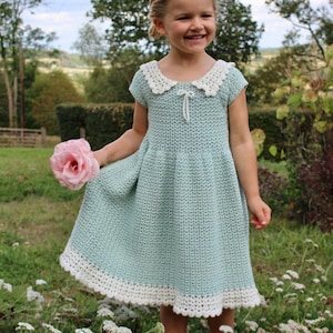 May include: A light sage green crochet dress featuring a white scalloped collar, short sleeves, and a white crocheted hem. The dress has a small white bow detail at the centre of the neckline and a textured, woven stitch pattern throughout the garment.