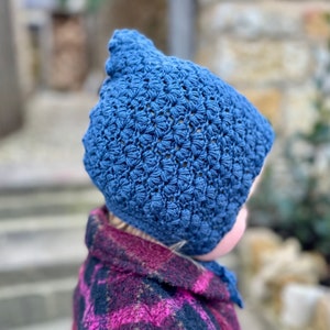 May include: A blue crocheted hat with a textured pattern. The hat is being worn by a person wearing a plaid shirt.