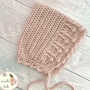 Puede incluir: Un gorro de bebé de ganchillo de color rosa claro con un borde festoneado y una cinta. El gorro tiene un diseño texturizado con pequeños bultos en relieve.