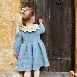May include: A young girl wearing a light blue crocheted dress with a white crocheted collar. She is standing in front of a wooden door with an old-fashioned door handle.