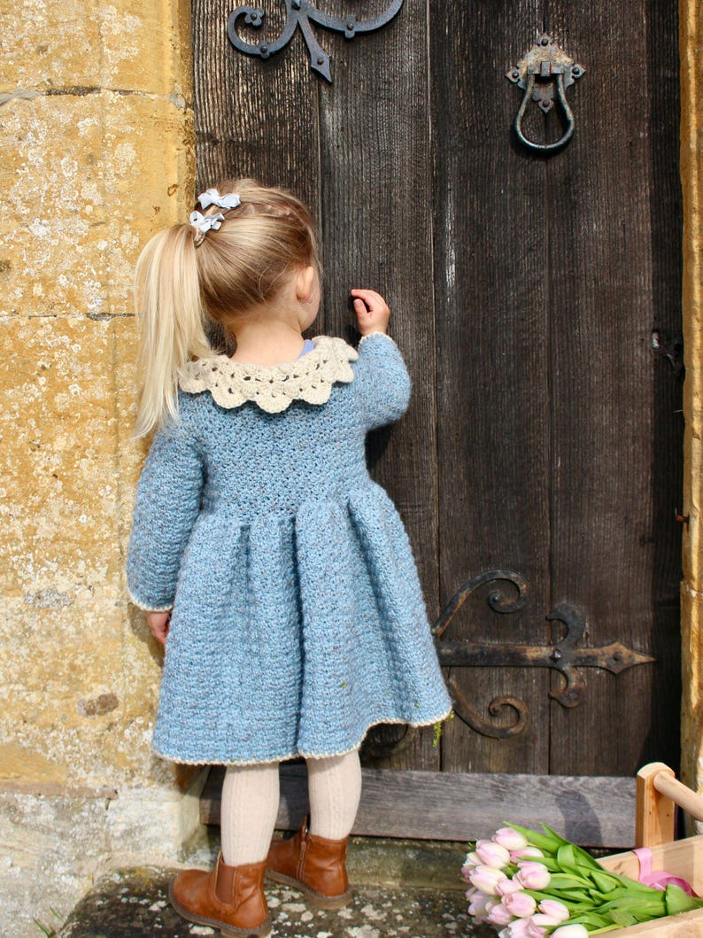 May include: A young girl wearing a light blue crocheted dress with a white crocheted collar. She is standing in front of a wooden door with an old-fashioned door handle.