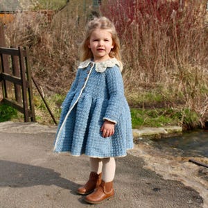 Könnte beinhalten: Ein junges Mädchen trägt ein hellblaues Strickkleid mit einem weißen Spitzenkragen. Sie steht auf einem gepflasterten Weg vor einem Steinhaus.