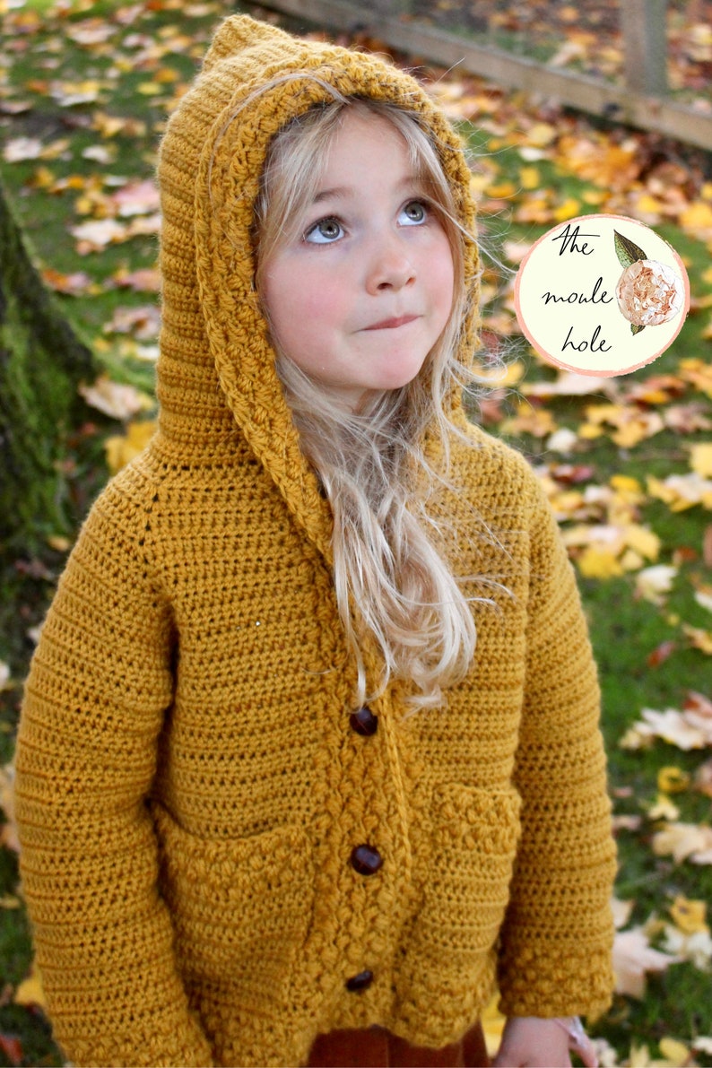 May include: A mustard yellow crocheted hooded cardigan with a button closure and pockets. The cardigan is made with a textured stitch and has a simple design.