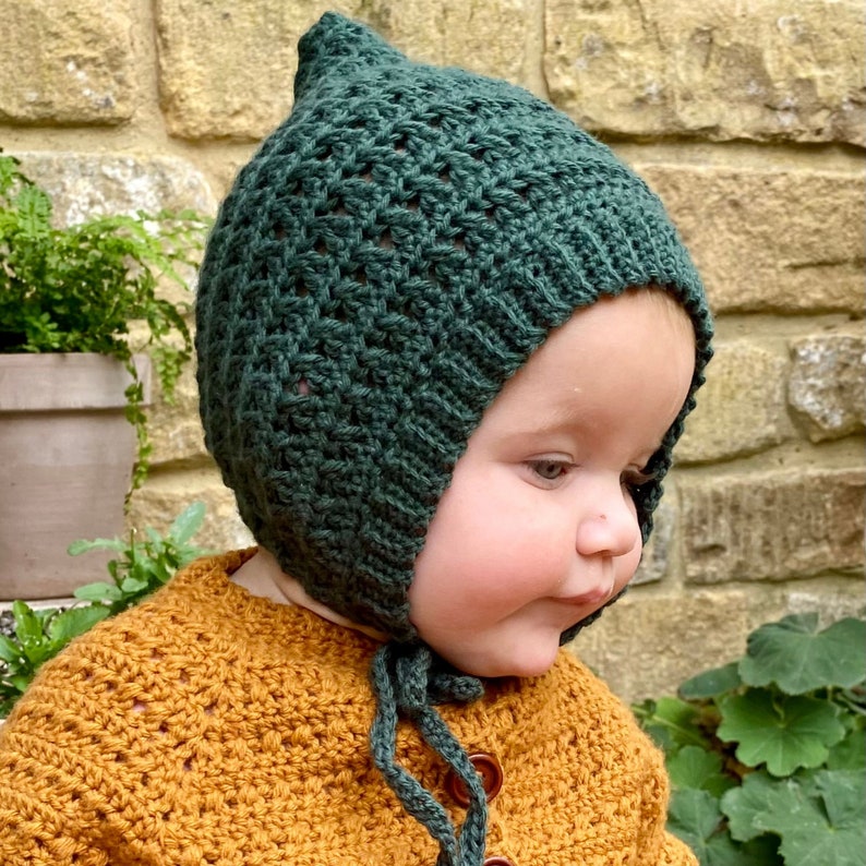Puede incluir: Un beb&eacute; con un gorro de punto verde con un lazo. El gorro tiene un patr&oacute;n de encaje y est&aacute; atado debajo de la barbilla. El beb&eacute; lleva un su&eacute;ter de punto amarillo.