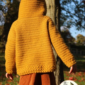May include: A mustard yellow crocheted hooded sweater with a ribbed texture. The sweater has long sleeves and a fitted silhouette. The sweater is worn over a brown skirt.