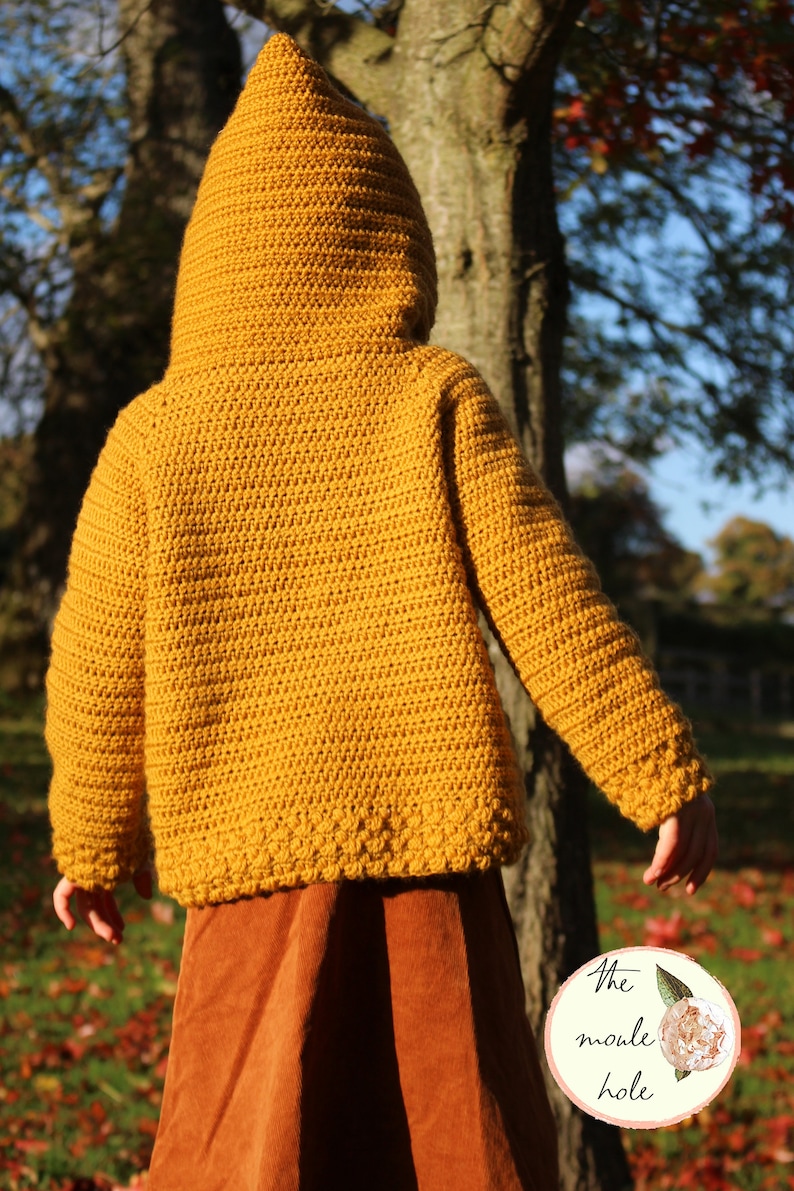 May include: A mustard yellow crocheted hooded sweater with a ribbed texture. The sweater has long sleeves and a fitted silhouette. The sweater is worn over a brown skirt.