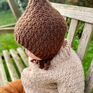 May include: A brown crocheted baby bonnet with a pointed top and a long tassel. The bonnet is worn by a child wearing a beige crocheted sweater and brown pants.
