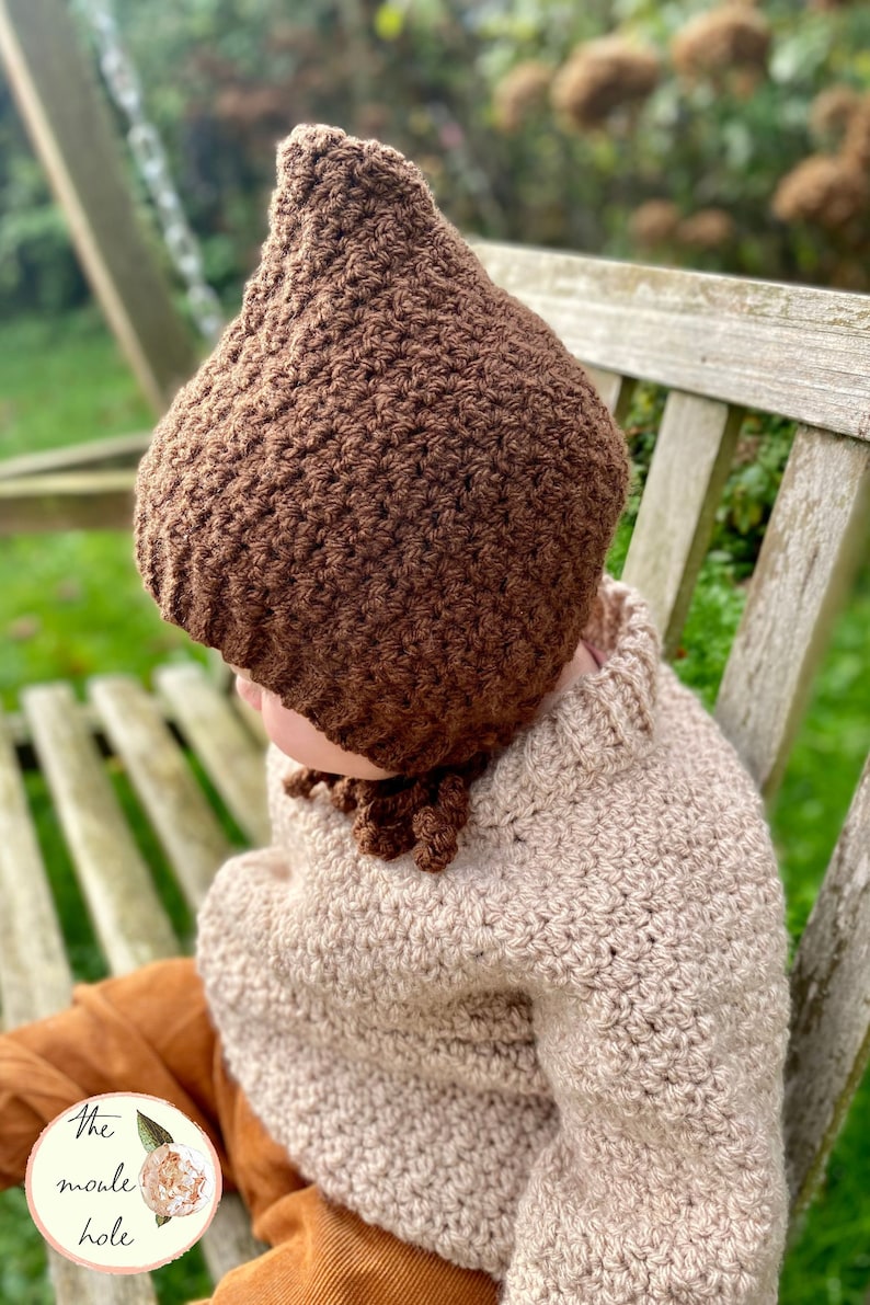 May include: A brown crocheted baby bonnet with a pointed top and a long tassel. The bonnet is worn by a child wearing a beige crocheted sweater and brown pants.