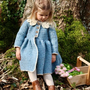 May include: A young girl wearing a blue crocheted coat with a white crocheted collar and brown boots. She is holding a bunch of pink tulips.