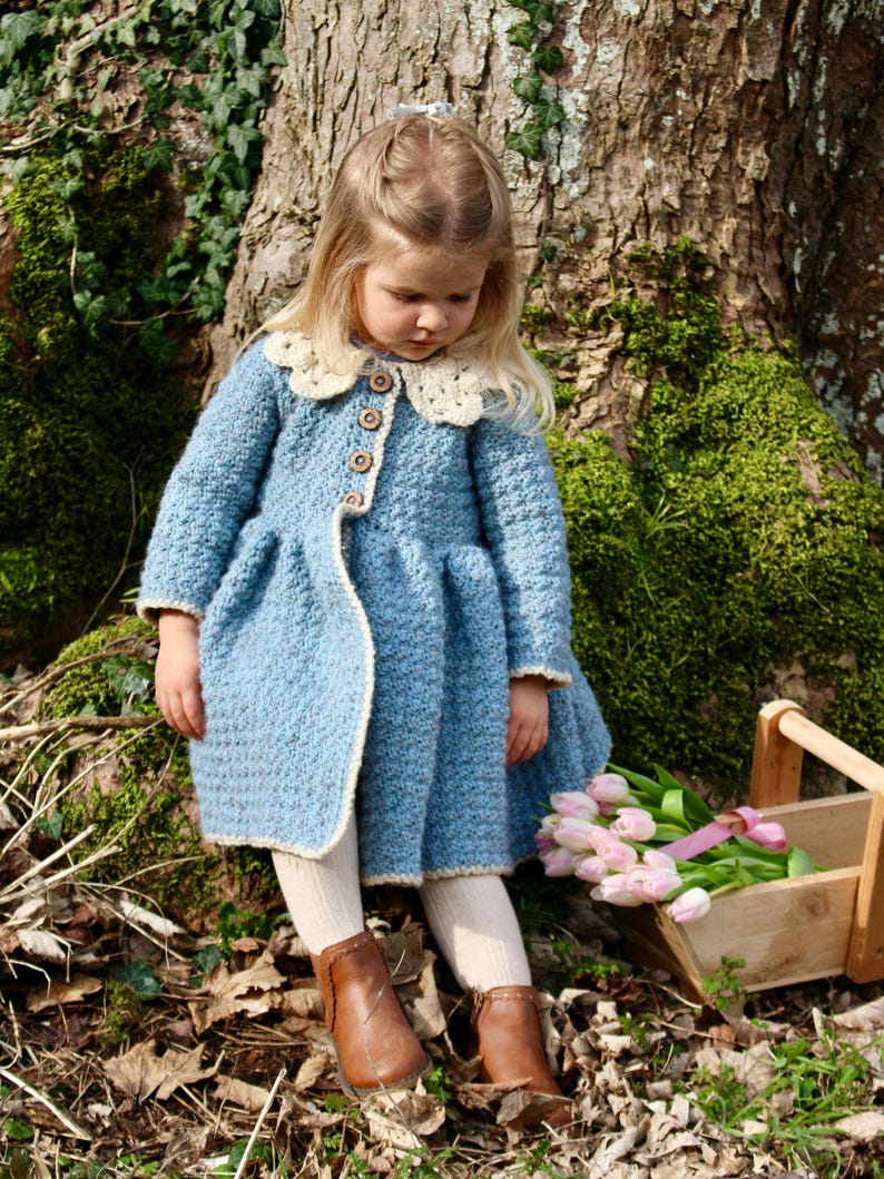 May include: A young girl wearing a blue crocheted coat with a white crocheted collar and brown boots. She is holding a bunch of pink tulips.