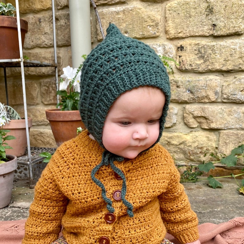 Puede incluir: Un beb&eacute; con un gorro de punto verde con la parte superior puntiaguda y un c&aacute;rdigan de punto marr&oacute;n con botones.