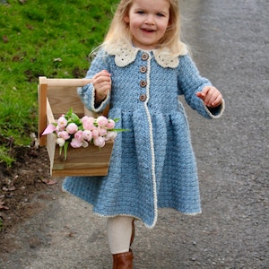 May include: A young girl wearing a light blue crocheted coat with a white crocheted collar and buttons. She is holding a wooden box filled with pink flowers.