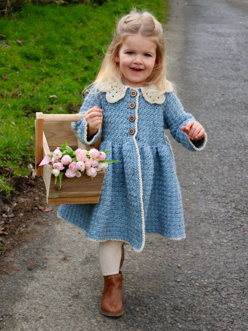 May include: A young girl wearing a light blue crocheted coat with a white crocheted collar and buttons. She is holding a wooden box filled with pink flowers.