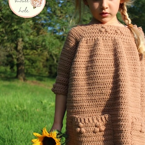 May include: A young girl wearing a brown crocheted dress with a pocket and holding a sunflower. The dress has a crocheted texture and a round neckline. The girl has blonde hair and is wearing a yellow shirt underneath the dress.
