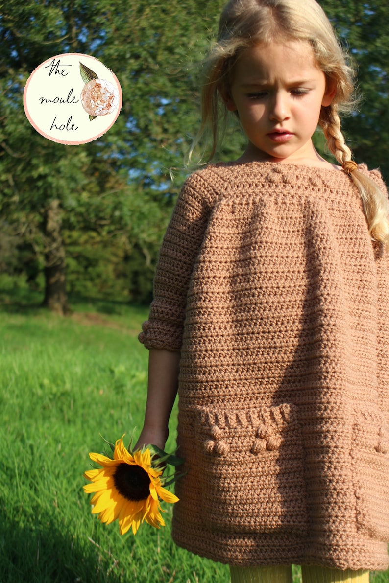 May include: A young girl wearing a brown crocheted dress with a pocket and holding a sunflower. The dress has a crocheted texture and a round neckline. The girl has blonde hair and is wearing a yellow shirt underneath the dress.
