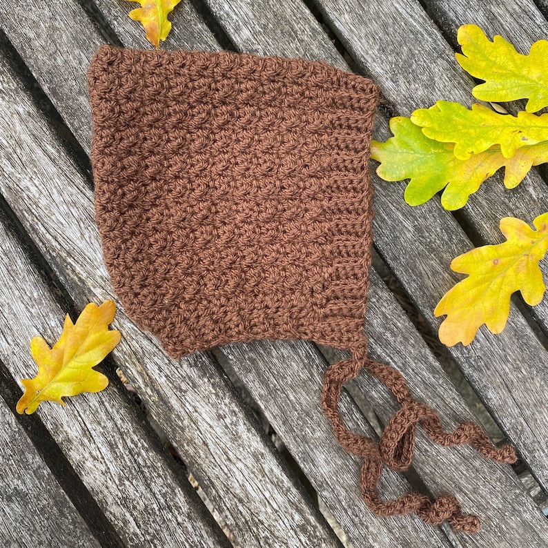 May include: A brown crocheted baby bonnet with a long tie. The bonnet is lying on a wooden surface with autumn leaves.