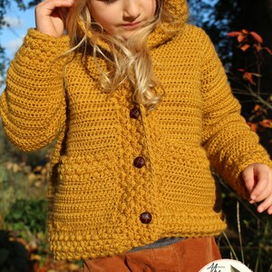 May include: A mustard yellow crocheted hooded cardigan with brown buttons. The cardigan is worn by a child and has a textured stitch pattern.