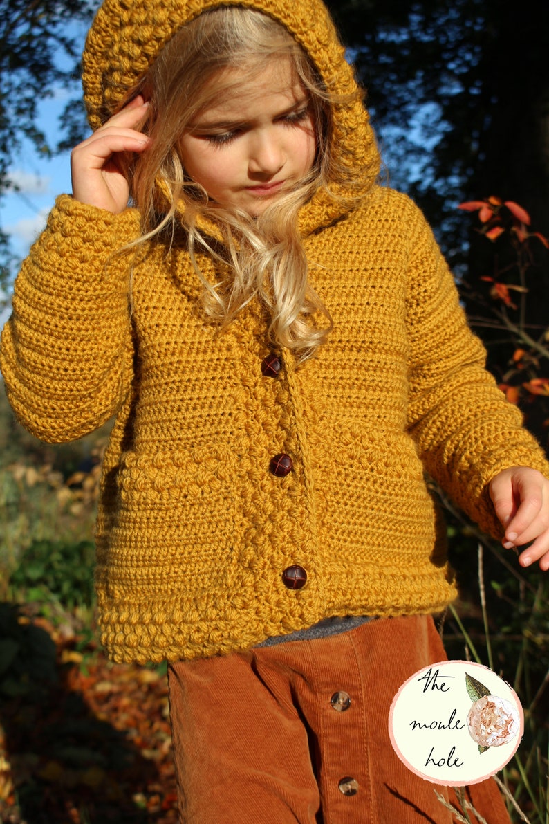 May include: A mustard yellow crocheted hooded cardigan with brown buttons. The cardigan is worn by a child and has a textured stitch pattern.