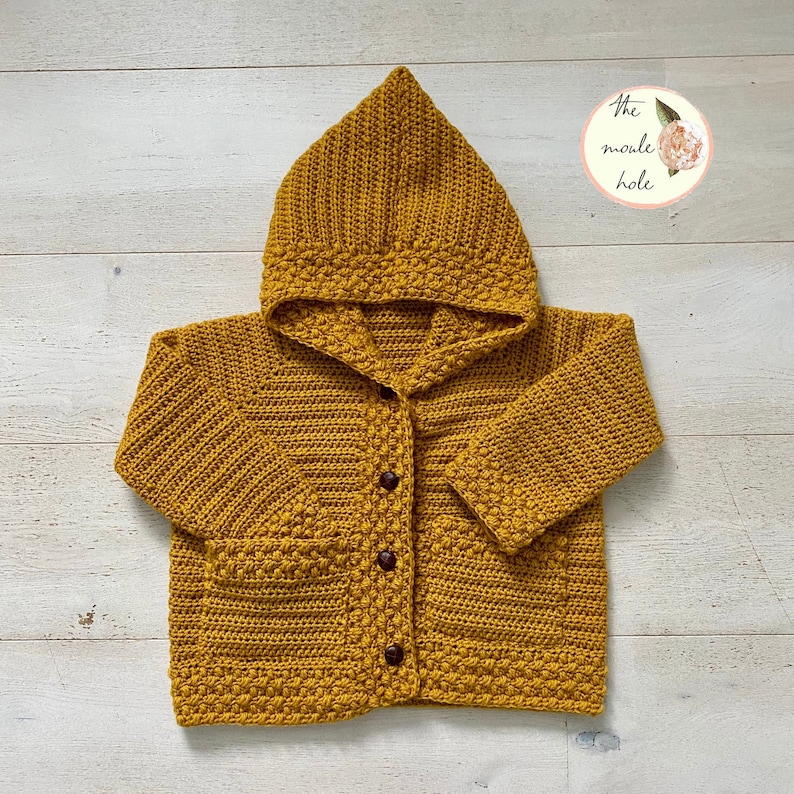 May include: A mustard yellow crocheted hooded cardigan with pockets and brown buttons. The cardigan is laid flat on a white wooden surface.