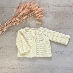 May include: A crocheted baby cardigan in a cream colour with a button fastening. The cardigan features a delicate crocheted pattern around the neckline and a textured stitch throughout.