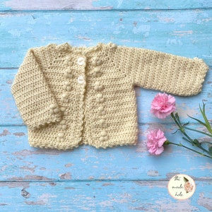 May include: A crocheted baby cardigan in a pale yellow colour with a button fastening. The cardigan has a textured stitch pattern and a scalloped edge. The cardigan is photographed against a blue wooden background with pink flowers.