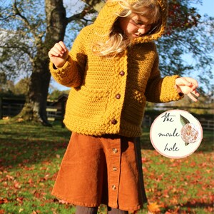 May include: A young person wearing a mustard yellow crocheted cardigan with a hood and matching corduroy skirt. The cardigan has a button closure and the skirt has a button-up front. The person is standing in a grassy area with fallen leaves.