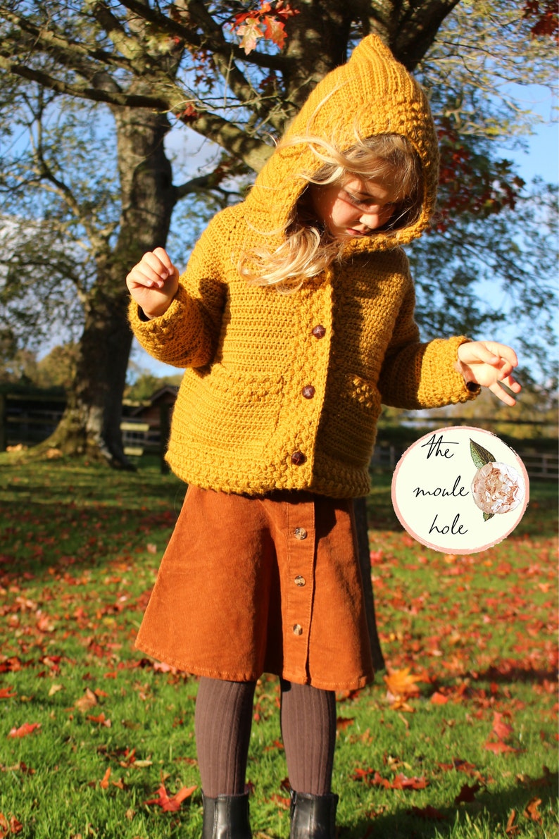 May include: A young person wearing a mustard yellow crocheted cardigan with a hood and matching corduroy skirt. The cardigan has a button closure and the skirt has a button-up front. The person is standing in a grassy area with fallen leaves.