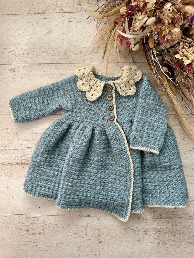 May include: A blue crocheted baby cardigan with a white crocheted collar and wooden buttons.