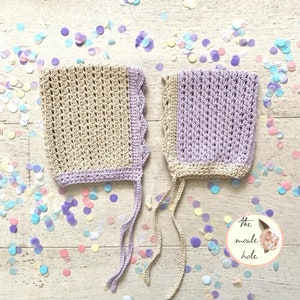 May include: Two crocheted baby bonnets, one in beige and one in lavender, with white ties. The bonnets are on a white wooden surface with confetti.