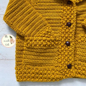 May include: A mustard yellow crocheted cardigan with a pocket and buttons. The cardigan is made with a textured stitch.