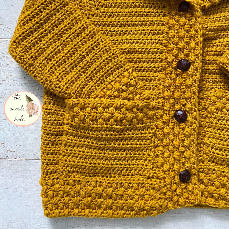 May include: A mustard yellow crocheted cardigan with a pocket and buttons. The cardigan is made with a textured stitch.