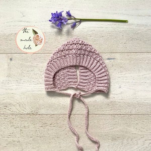 Peut inclure: Un bonnet de bébé en crochet rose avec un lien à l'arrière. Le bonnet est sur un fond en bois clair. Le texte "The noodle hole" est visible sur l'image.